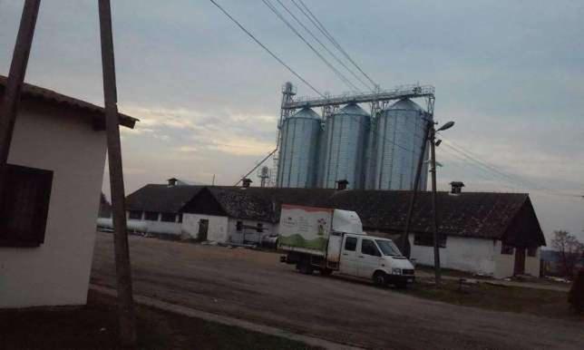 Metalowe silosy na tle budynków gospodarczych i szarego nieba. Na pierwszym planie droga i biały samochód dostawczy z reklamą.