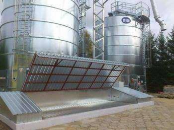 Silosy zbożowe z metalu, otwarte zadaszenie. Widoczne drabiny i rury. Tło stanowią drzewa iglaste. Ujęcie z dołu, podkreślające wielkość zbiorników.