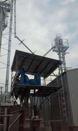 Wysoka, zewnętrzna konstrukcja stalowa z platformą i niebieskim urządzeniem, widoczna z dołu na tle pochmurnego nieba, element budowy hali.