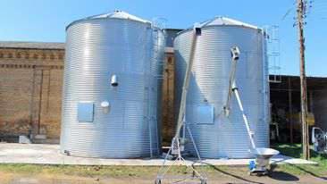 Dwa srebrne silosy zbożowe z metalowymi schodkami i podajnikiem ślimakowym na tle ceglanego budynku i błękitnego nieba. Widok z poziomu gruntu, oświetlenie dzienne.