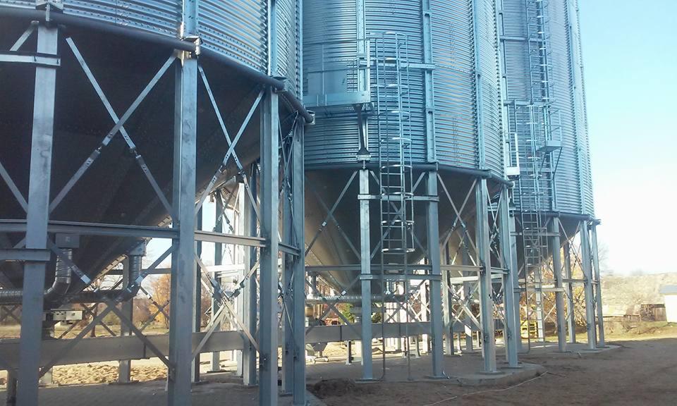 Silosy zbożowe na stalowej konstrukcji wsporczej z drabinkami, widok z dołu, błękitne niebo w tle. Konstrukcja lekka, ażurowa, ocynkowana.