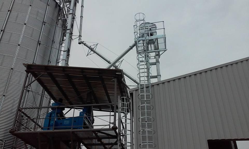 Silos zbożowy z metalową konstrukcją schodów i podestem, połączony z budynkiem o metalowej elewacji, na tle pochmurnego nieba.