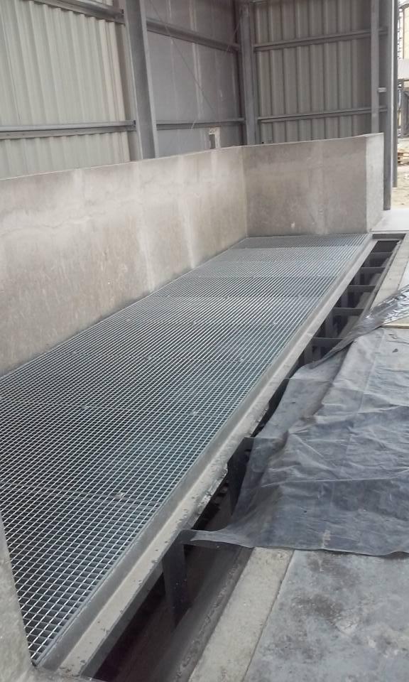 Zastosowanie kraty pomostowej w hali przemysłowej. Stalowa konstrukcja z betonowym obramowaniem. Widoczne elementy konstrukcyjne i czarna folia zabezpieczająca.