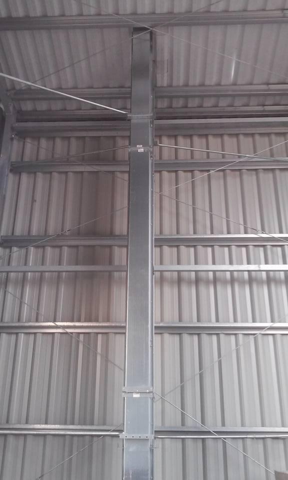 Wnętrze hali: stalowa kolumna nośna z widocznymi łączeniami, blaszane ściany i dach z widocznymi elementami konstrukcyjnymi. Szara kolorystyka, surowy industrialny klimat.