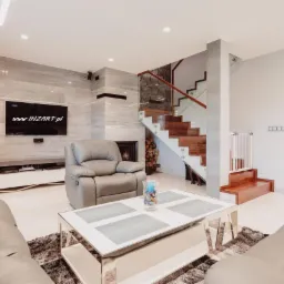 Jasne wnętrze salonu z marmurową ścianą, kominkiem, schodami z drewnianymi stopniami i szklaną balustradą, skórzanymi sofami i fotelem z funkcją relaks, niski stolik kawowy z metalowymi nogami...