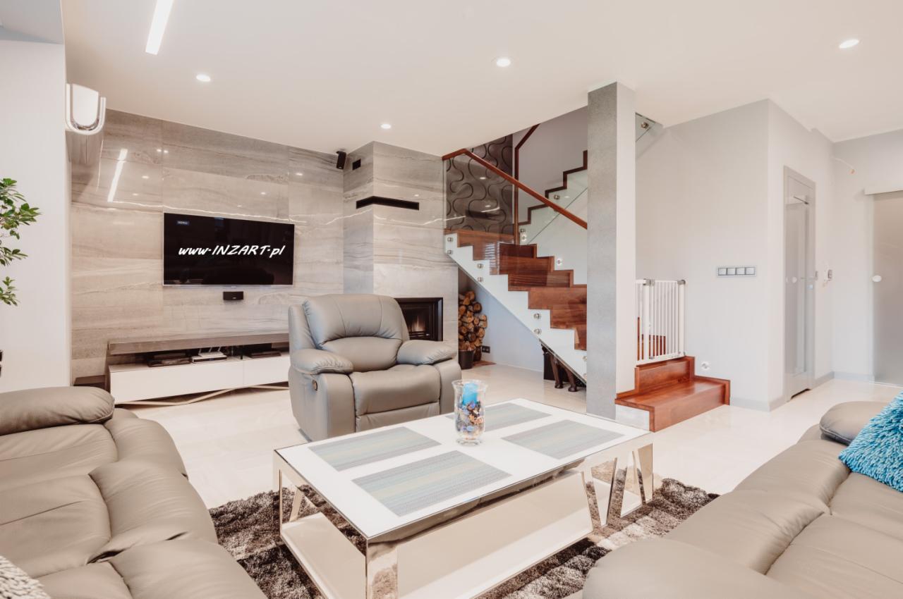 Jasne wnętrze salonu z marmurową ścianą, kominkiem, schodami z drewnianymi stopniami i szklaną balustradą, skórzanymi sofami i fotelem z funkcją relaks, niski stolik kawowy z metalowymi nogami...