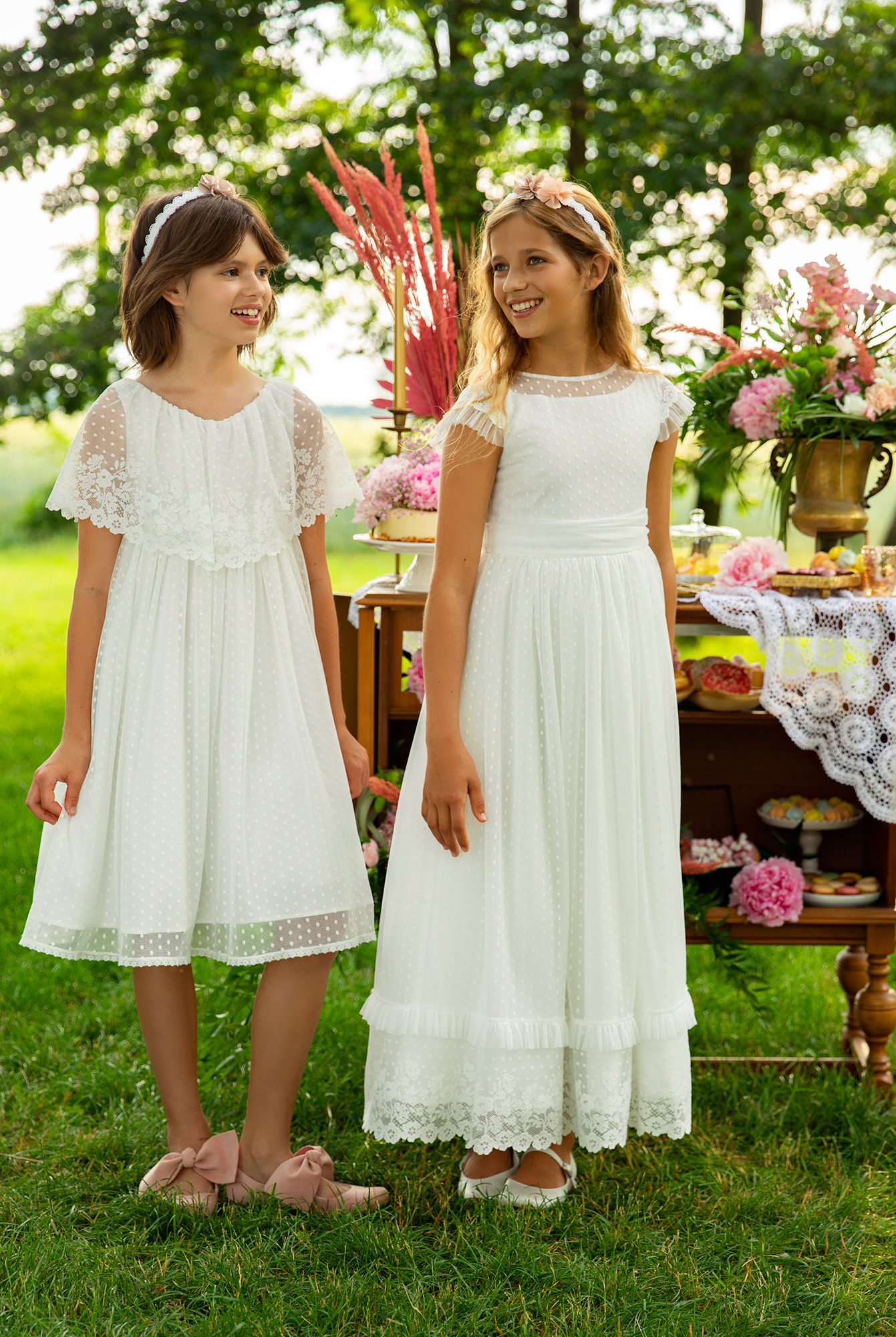 Dwie uśmiechnięte dziewczynki w białych, eleganckich sukienkach z koronkowymi detalami, stojące na trawie przed stołem z dekoracjami i słodkościami.