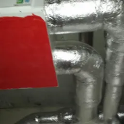 Izolowane, srebrną folią rury wentylacyjne, z podłączoną białą jednostką, widoczne przy szarej ścianie i suficie, w pomieszczeniu o słabym oświetleniu.