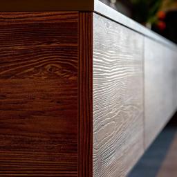 Rena Sp. z o.o. - Fragment szafki z frontami w dwóch odcieniach drewna: ciemnobrązowym i jasnoszarym z wyraźnym usłojeniem, widoczne kwiaty w tle.