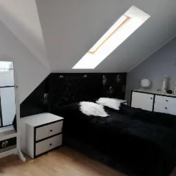 Sypialnia na poddaszu z czarnym łóżkiem o pluszowej teksturze, białymi poduszkami, szafką nocną i toaletką, oświetlona oknem dachowym.