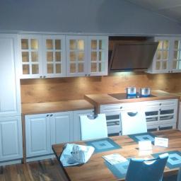 Rena Sp. z o.o. - Jasna kuchnia z białymi szafkami z przeszklonymi frontami, drewnianym blatem i brązową ścianą nad blatem, widoczny stół z krzesłami i garnki na płycie grzewczej.