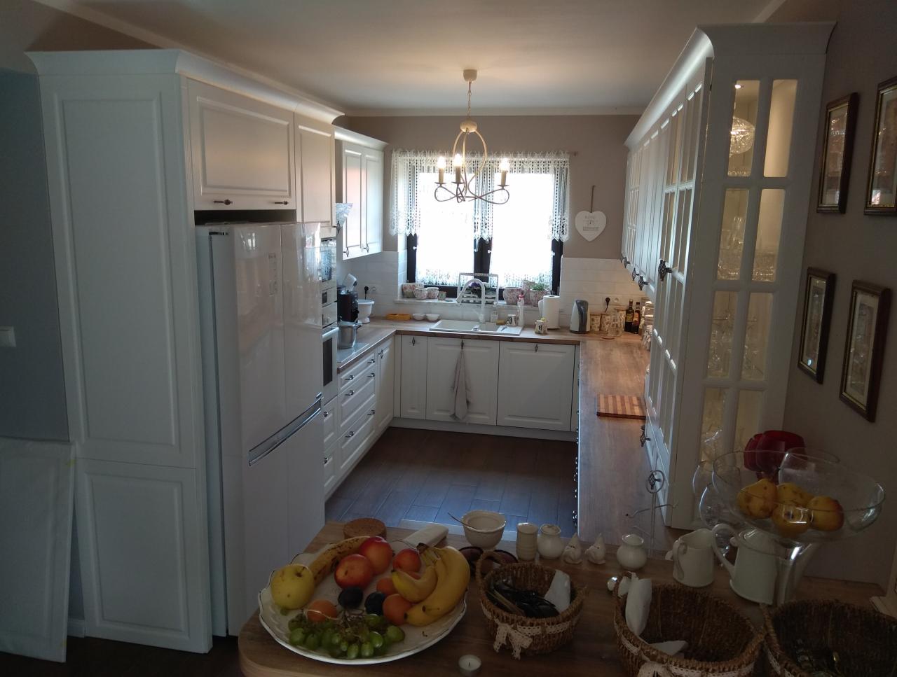 Jasna kuchnia z białymi meblami, drewnianym blatem i owocami na stole, widok na okno z firankami.