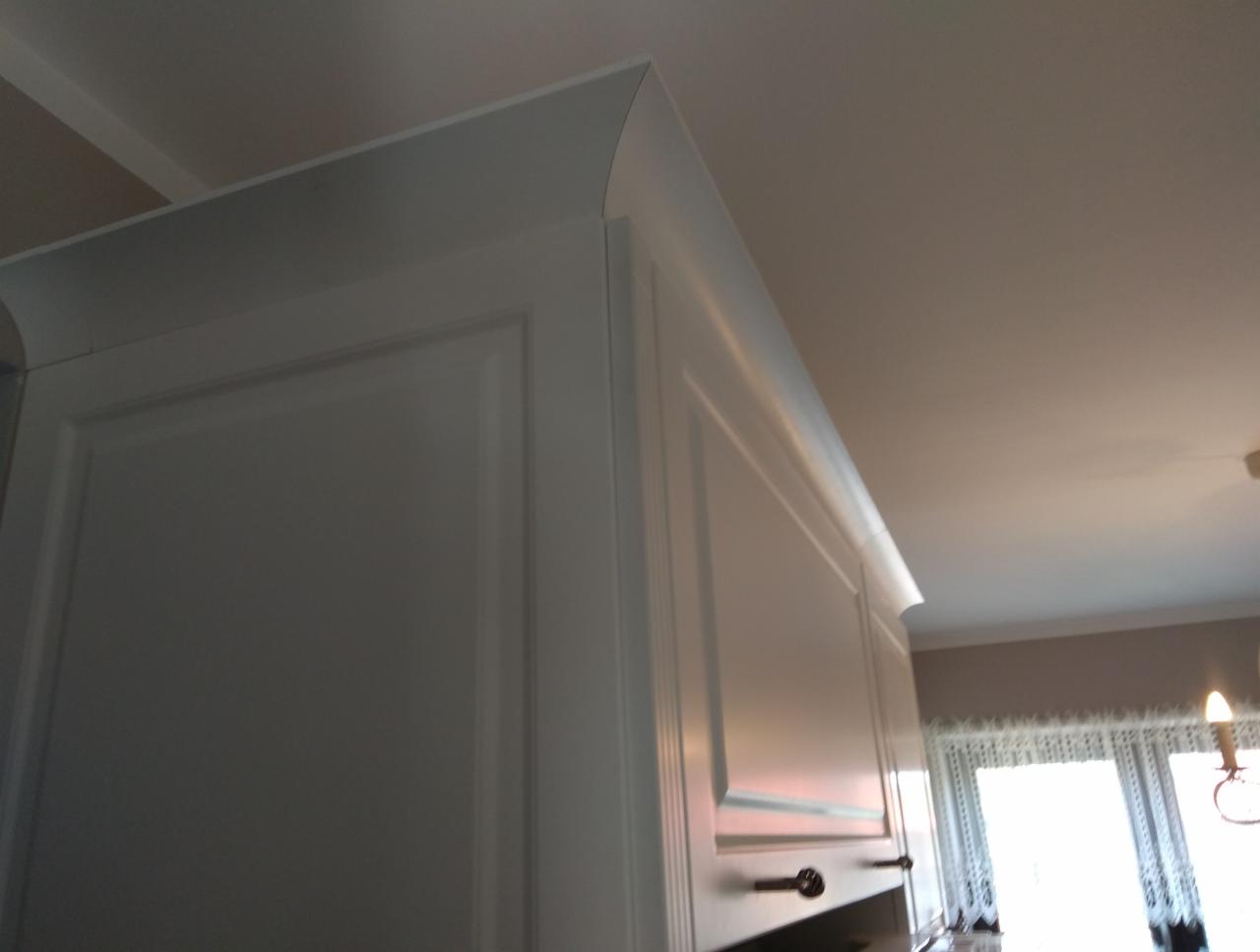 Fragment białej szafki kuchennej wiszącej, z ozdobnym frezowaniem i zaokrągloną górną krawędzią, widok z dołu w kierunku sufitu i okna.