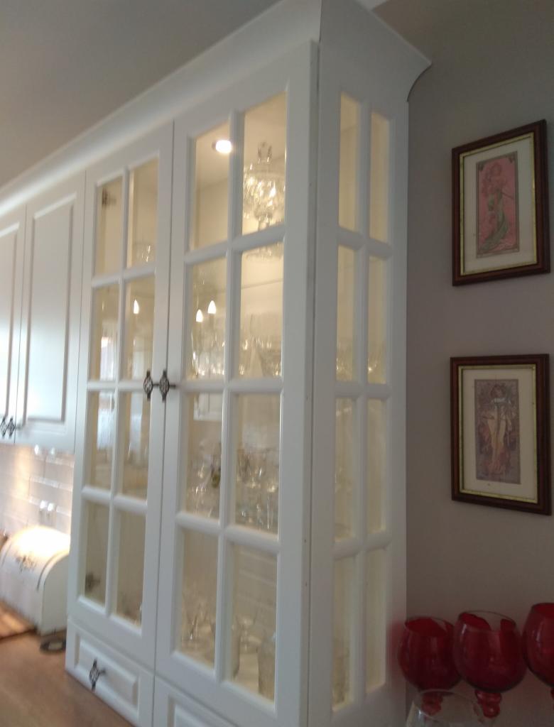 Biała, przeszklona szafka kuchenna z dekoracyjnymi uchwytami, wypełniona szkłem i kryształami, oświetlona od wewnątrz, z obrazkami w ramkach na ścianie obok.