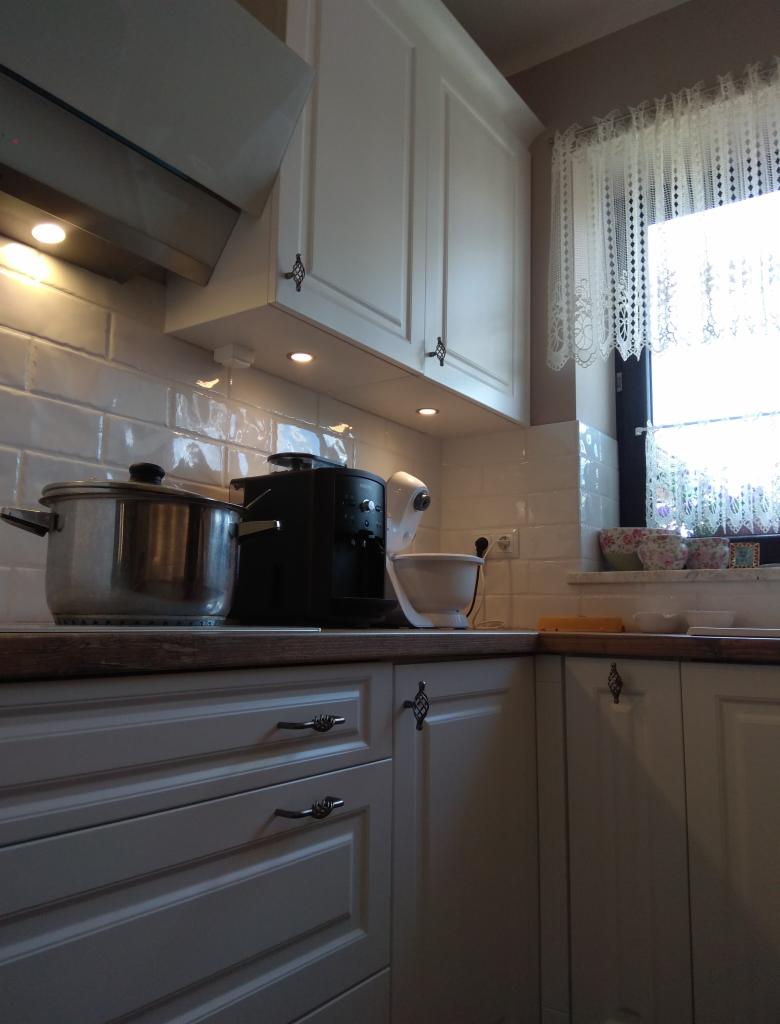 Fragment kuchni z białymi, frezowanymi frontami szafek z ozdobnymi uchwytami, widoczny garnek na płycie, ekspres do kawy i mikser, białe płytki na ścianie i okno z koronkową firanką.