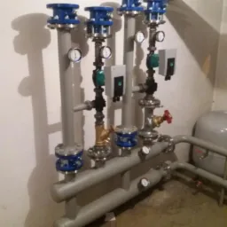 Szare rury instalacji grzewczej z manometrami, zaworami i niebieskimi elementami łączeniowymi na tle białej ściany.