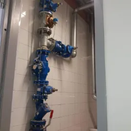 Pionowa instalacja wodna z niebieskimi zaworami i srebrnymi rurami zamontowana na ścianie z białych kafelków, widok z dołu.