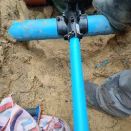 Niebieska rura wodociągowa z czarnym zaworem łączącym, zakopana w piasku, obok widoczne fragmenty ubrań roboczych i ręcznik.