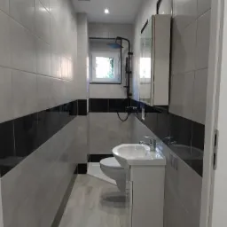 Wąska łazienka po remoncie z szarymi i czarnymi płytkami, białą umywalką z szafką, toaletą i kabiną prysznicową z czarną armaturą.