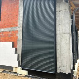 P.H.U FASMONT-GRUPA - Zamknięte, grafitowe żaluzje fasadowe na surowej elewacji domu w trakcie budowy. Widoczne detale montażu i ocieplenie budynku.