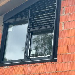 P.H.U FASMONT-GRUPA - Zamontowane czarne żaluzje fasadowe na oknie w surowym murze z cegły. Widoczne elementy montażowe i konstrukcja okna. Kątowe ujęcie z dołu.