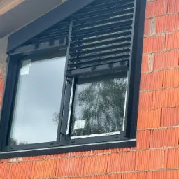 Zamontowane czarne żaluzje fasadowe na oknie w surowym murze z cegły. Widoczne elementy montażowe i konstrukcja okna. Kątowe ujęcie z dołu.