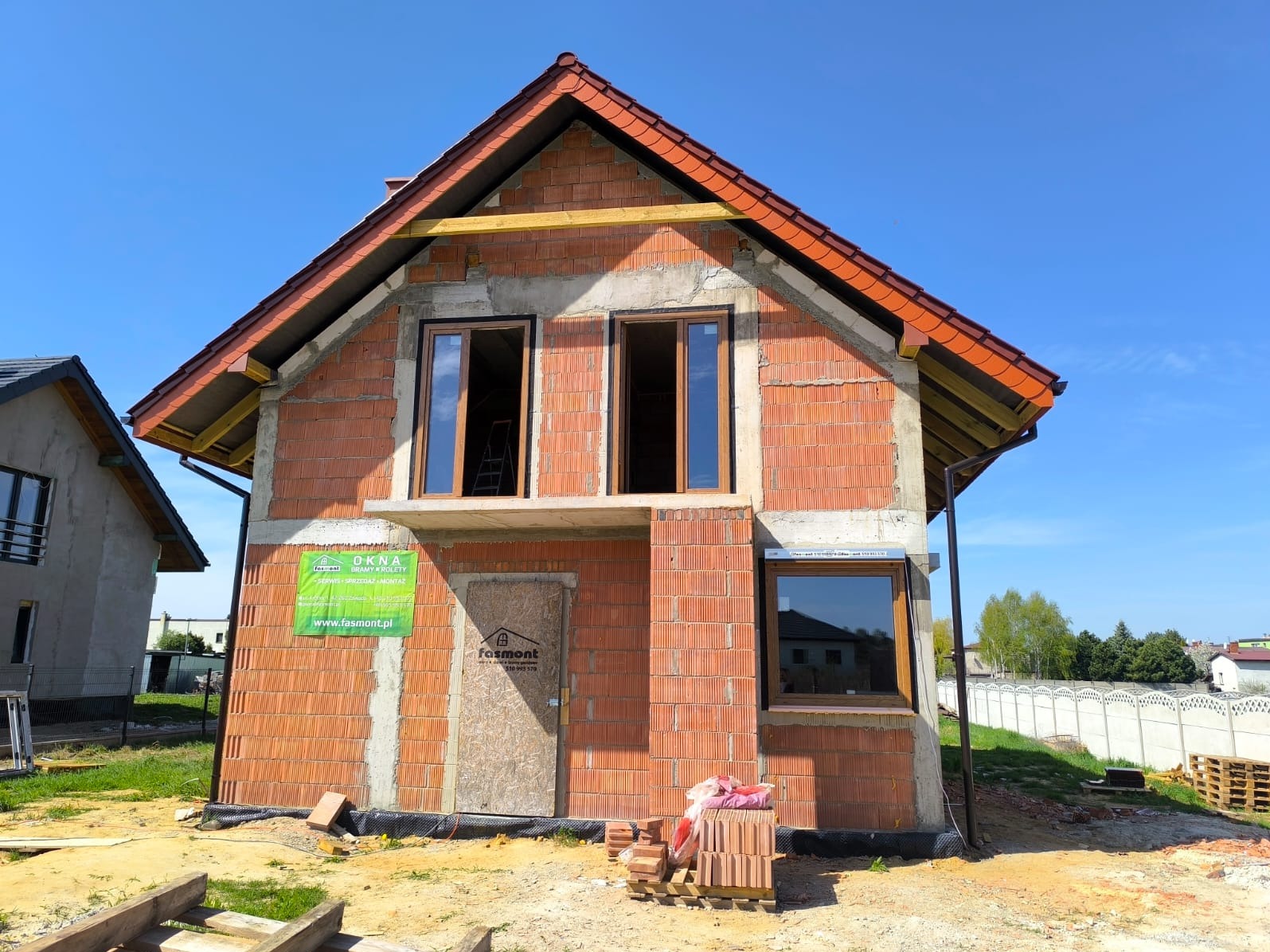 Surowy dom z czerwonej cegły z zamontowanymi oknami z brązowymi ramami i zieloną reklamą firmy. W tle błękitne niebo i częściowo widoczny drugi dom.