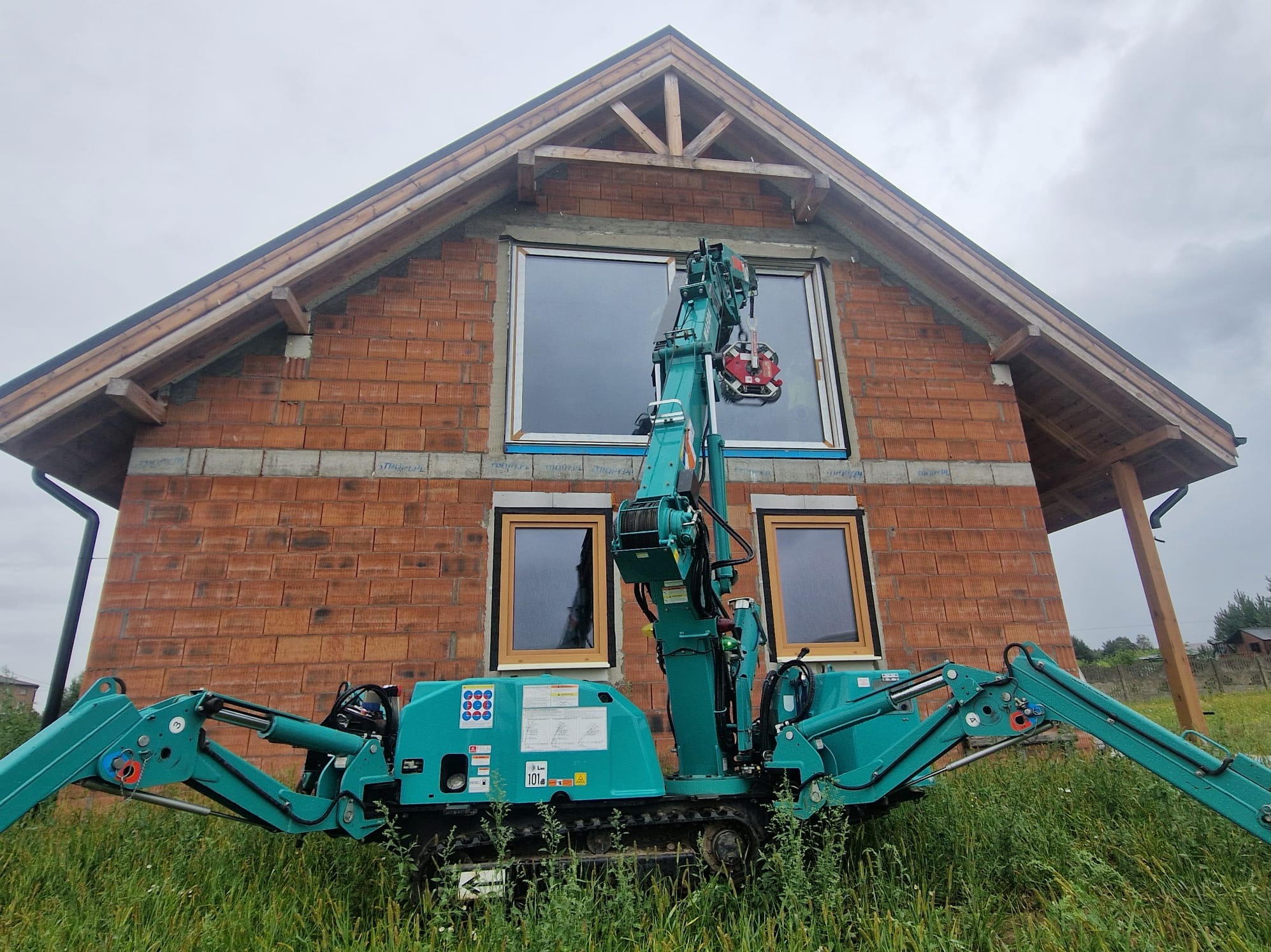 Montaż okna w surowym domu z cegły za pomocą specjalistycznego dźwigu, widok z dołu. Dom w trakcie budowy, okna z ramami w kolorze drewna.