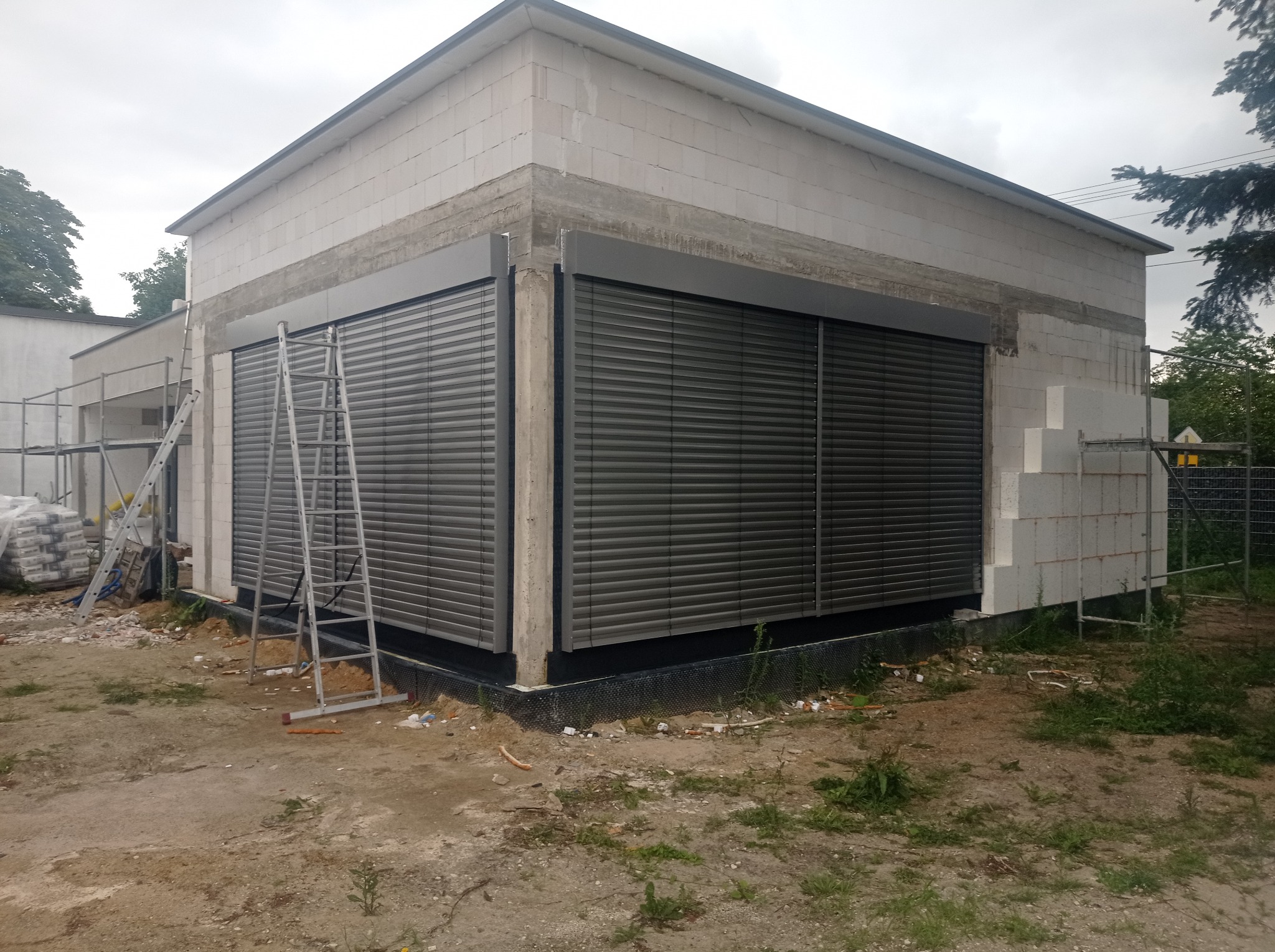 Budynek w trakcie budowy z zamontowanymi żaluzjami fasadowymi w kolorze grafitowym, z drabiny opartą o ścianę. Widoczne materiały budowlane i rusztowanie.