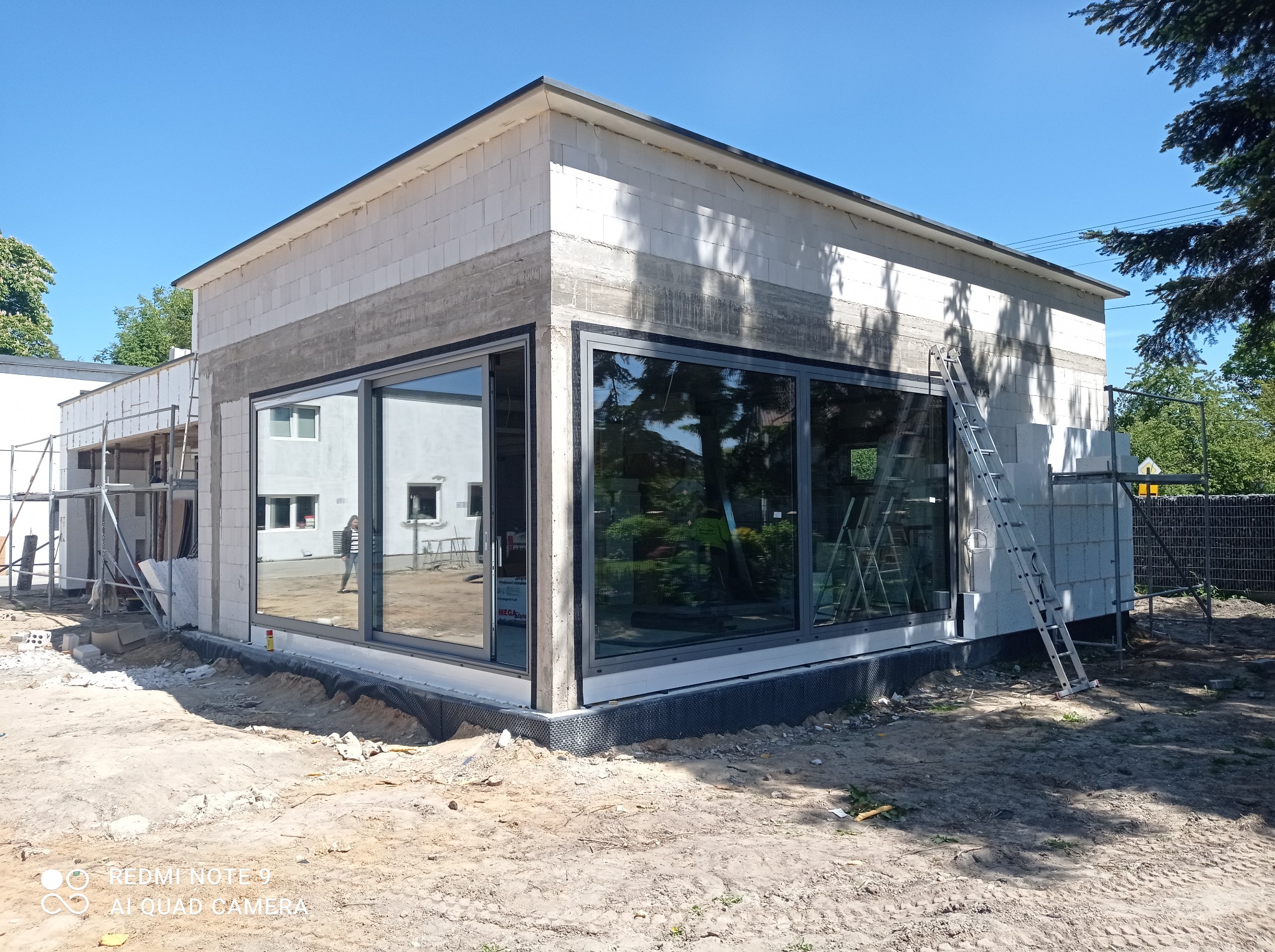 Surowy budynek z bloków z dużymi oknami w szarych ramach, drabina oparta o ścianę, ziemia wokół, widok na sąsiedni budynek i drzewa, słoneczny dzień.