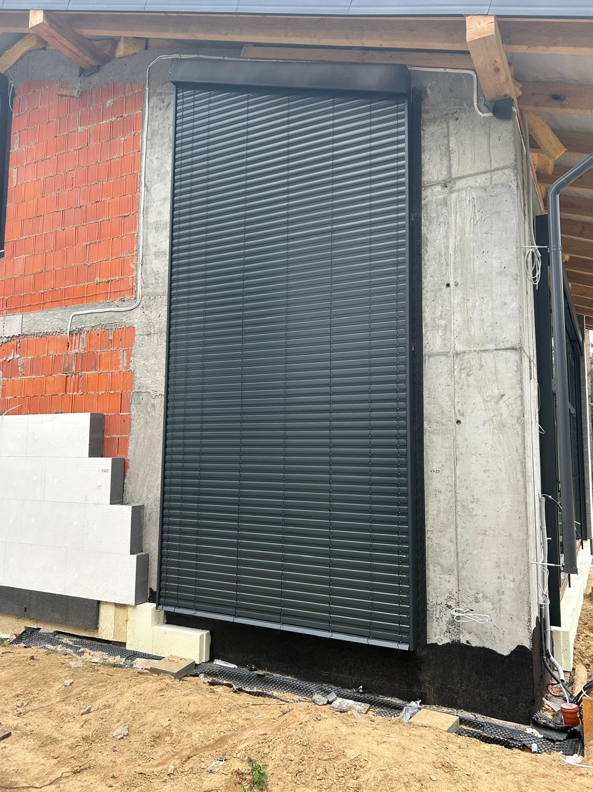 Zamknięte, grafitowe żaluzje fasadowe na surowej elewacji domu w trakcie budowy. Widoczne detale montażu i ocieplenie budynku.