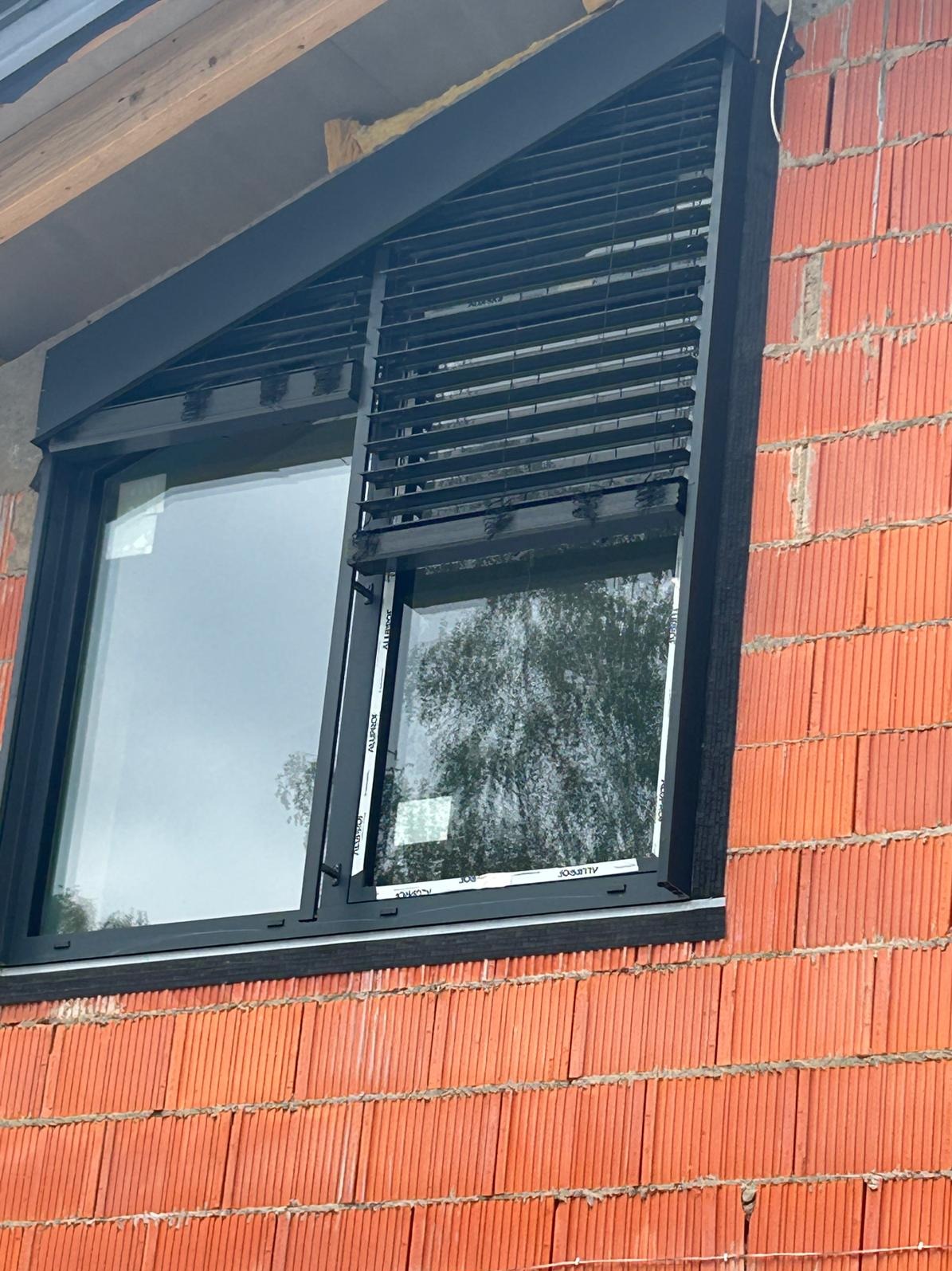 Zamontowane czarne żaluzje fasadowe na oknie w surowym murze z cegły. Widoczne elementy montażowe i konstrukcja okna. Kątowe ujęcie z dołu.