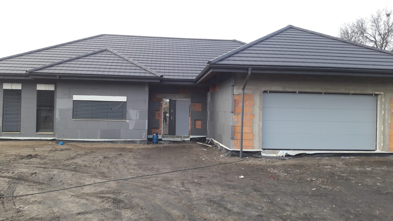 Surowy stan budowy domu jednorodzinnego z szarej cegły, widoczne zamontowane żaluzje zewnętrzne i segmentowa brama garażowa w kolorze szarym.