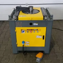 Żółto-szara giętarka do stali zbrojeniowej z pedałem nożnym, widok z przodu na tle białej bramy garażowej. Sprzęt do obróbki stali.