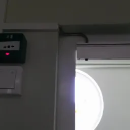 Zielony przycisk alarmowy z czerwoną diodą i strzałkami, zamontowany obok wyłącznika światła, w pobliżu automatycznego otwierania drzwi z widoczną rurką ochronną kabla.