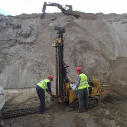 Badania geotechniczne gruntu: dwóch geologów w kaskach i kamizelkach przy wiertnicy na tle piaszczystego wzgórza i koparki. Próbki gleby i samochód dostawczy.