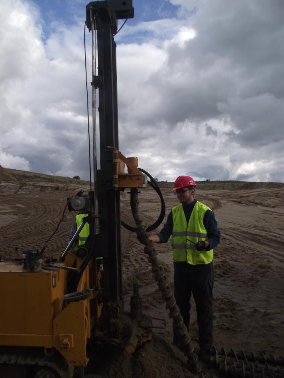 Pracownik w kasku i kamizelce obsługuje wiertnicę na tle piaszczystego terenu i pochmurnego nieba. Wiertło zagłębione w ziemi.