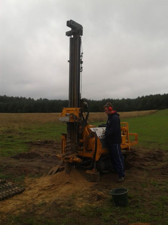 Żółta wiertnica geologiczna w akcji na polu, obsługiwana przez mężczyznę w słuchawkach. W tle las i pochmurne niebo. Widoczne wywiercone próbki gruntu.
