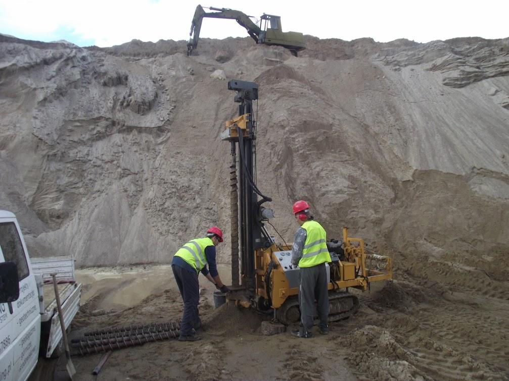 Badania geotechniczne gruntu: dwóch geologów w kaskach i kamizelkach przy wiertnicy na tle piaszczystego wzgórza i koparki. Próbki gleby i samochód dostawczy.