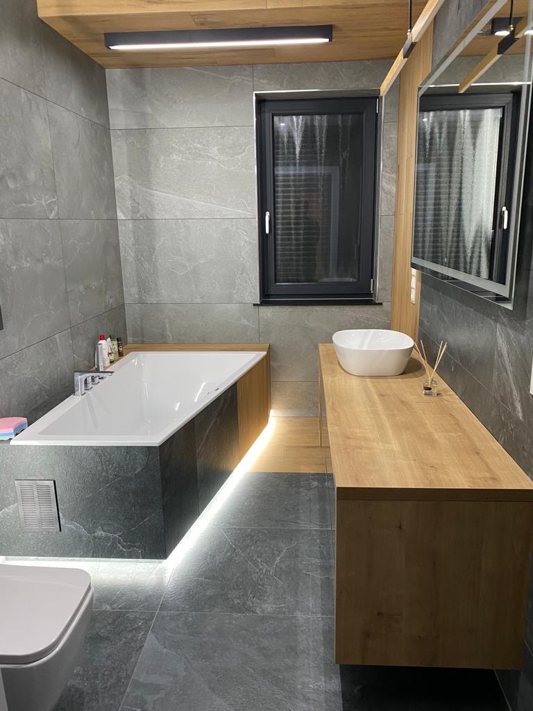 Nowoczesna łazienka z szarymi płytkami imitującymi marmur, podświetlana wanna obudowana ciemnym kamieniem, drewniana szafka z umywalką nablatową i czarne okno.