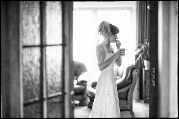 Czarno-białe zdjęcie panny młodej w sukni, z welonem, pijącej kawę w pokoju. Ujęcie przez otwarte drzwi, z widocznym fragmentem okna w tle.