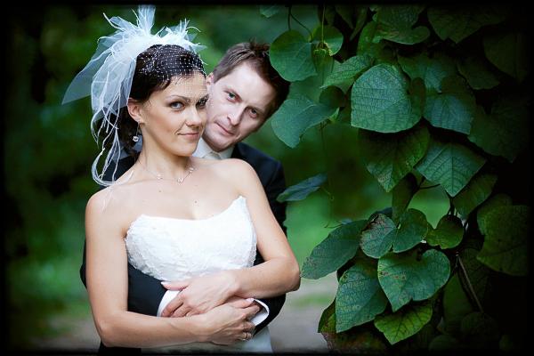 Uśmiechnięta panna młoda w białej sukni, obejmowana przez pana młodego na tle zielonej roślinności. Romantyczna fotografia ślubna w plenerze.