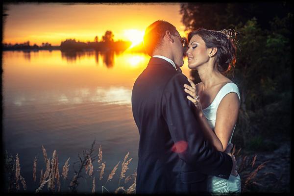 Para młoda w objęciach na tle jeziora o zachodzie słońca. Pan młody w ciemnym garniturze, panna młoda w białej sukni. Romantyczna sceneria, ciepłe kolory.