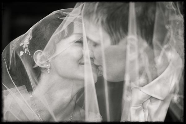 Czarno-białe zdjęcie pary młodej całującej się pod welonem. Widoczne detale: kolczyki panny młodej i wyraz twarzy.