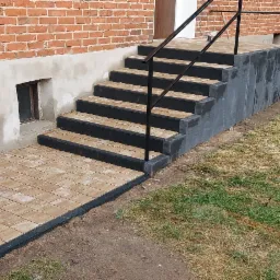 Wykończone schody wejściowe z kostki brukowej w odcieniach beżu i czerni, z czarną metalową barierką, prowadzące do budynku z czerwonej cegły z oknem piwnicznym, obok zaparkowany biały samochód...