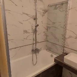 Wykończona łazienka z wanną i prysznicem typu deszczownica, marmurowe płytki na ścianach i podłodze, widoczne niedokończone punkty oświetleniowe na suficie i ścianie.