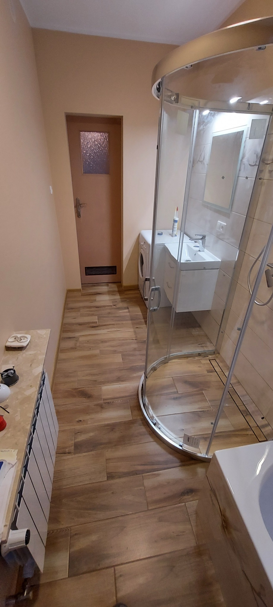 Nowoczesna łazienka z kabiną prysznicową typu walk-in, umywalką z szafką, pralką i grzejnikiem, wykończona płytkami imitującymi drewno.