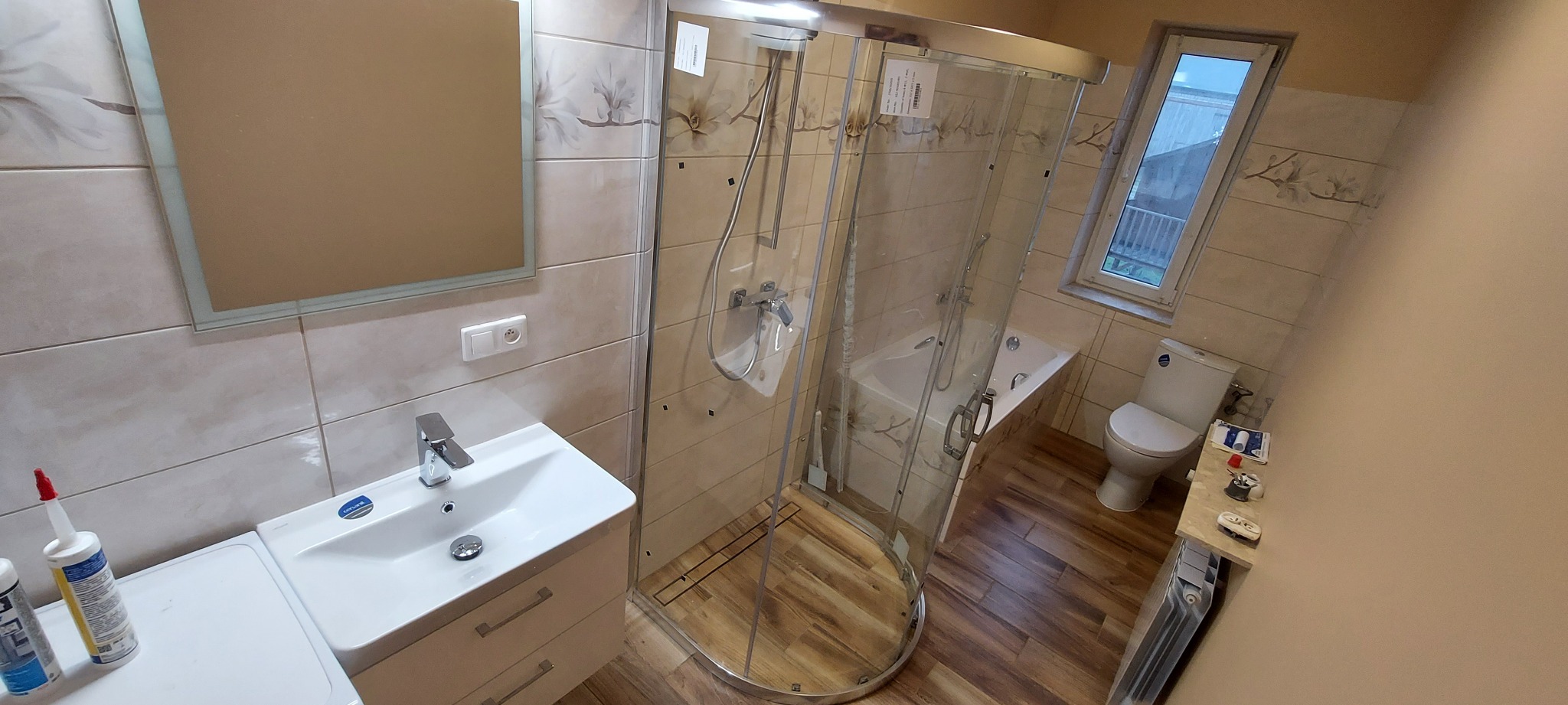 Ukończona łazienka z kabiną prysznicową z hydromasażem, białą umywalką z szafką, lustrem, toaletą i grzejnikiem, wykończona płytkami w beżowym odcieniu z motywem kwiatowym.