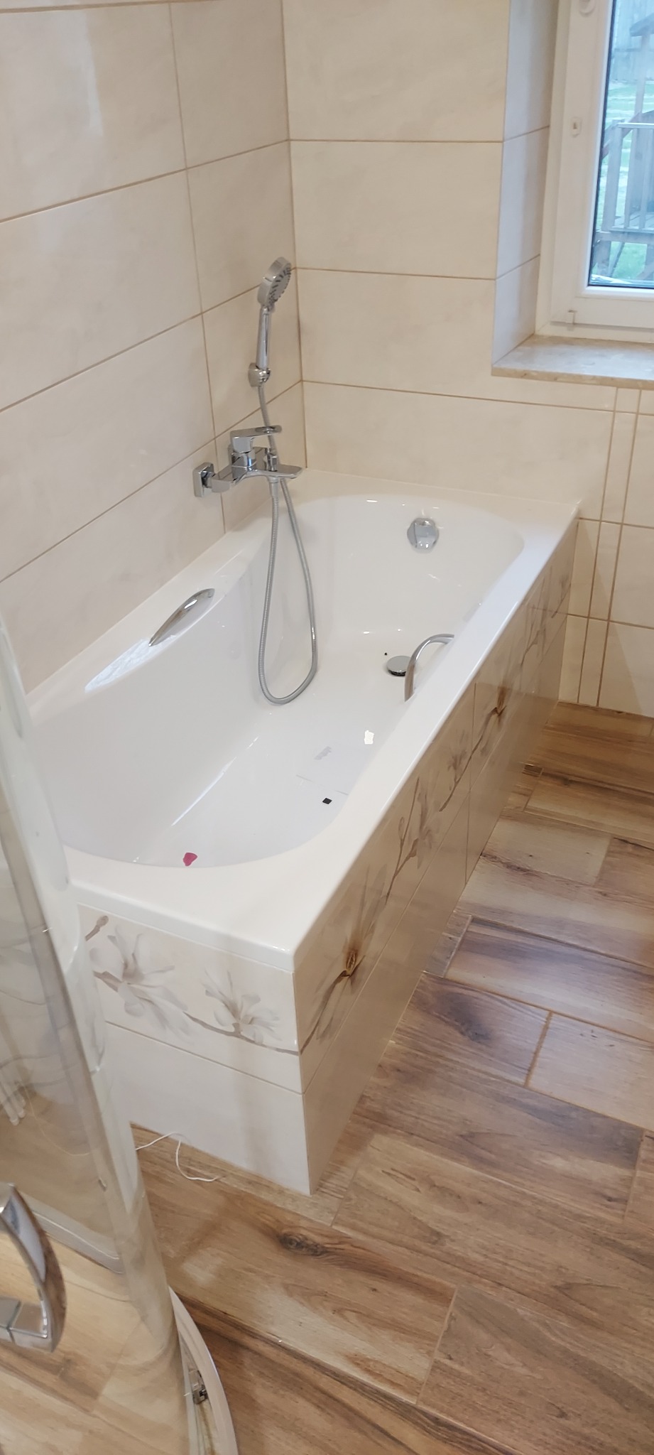 Biała wanna obudowana płytkami z motywem kwiatowym, chromowana bateria z prysznicem, beżowe płytki na ścianie i drewnopodobne płytki na podłodze w wykończonej łazience.