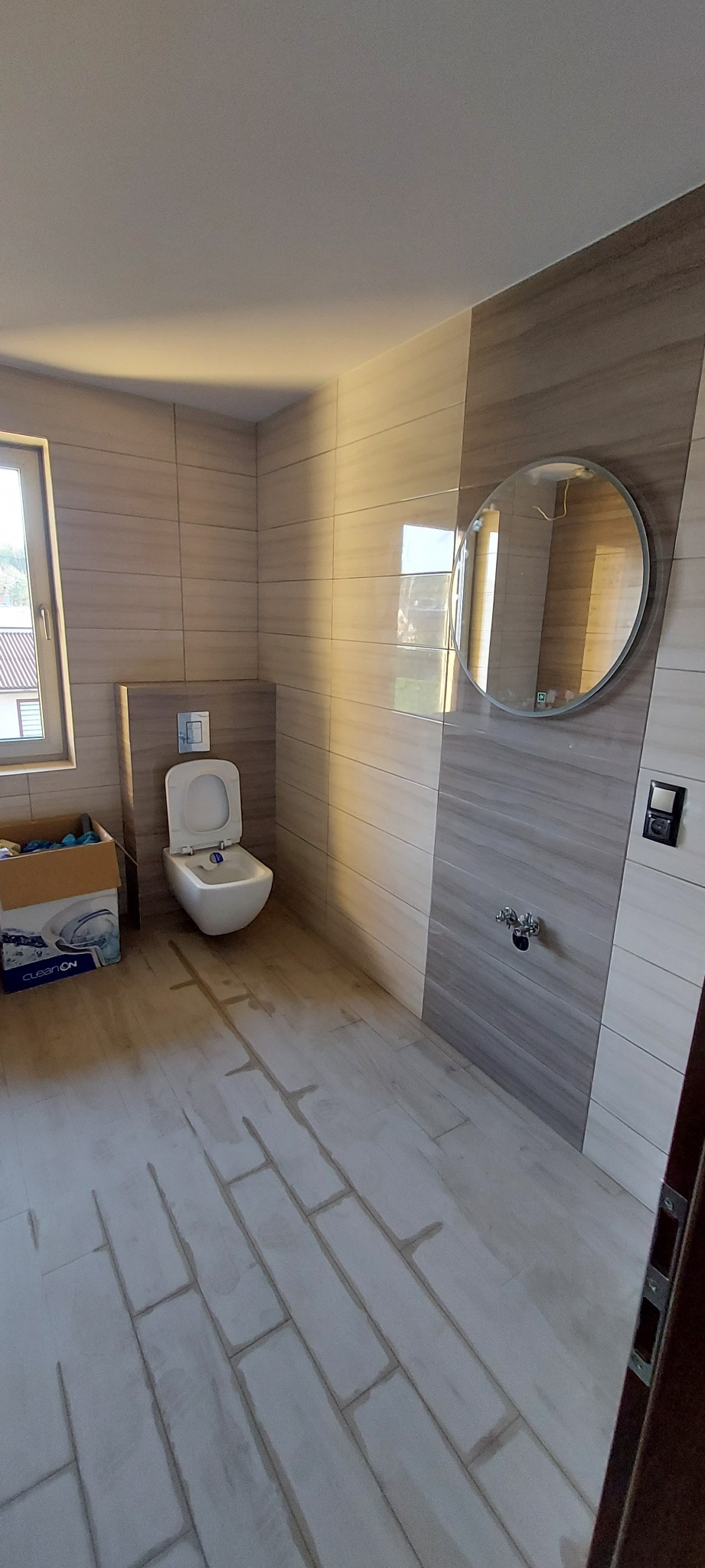 Nowa łazienka w trakcie wykańczania, widoczne płytki na ścianach w odcieniach szarości i beżu, biała toaleta podwieszana, okrągłe lustro i karton z wyposażeniem w rogu pomieszczenia.